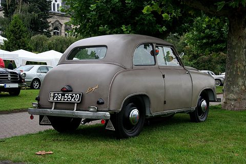 Rear of a small brown-grey two-door saloon with Lloyd script and mud flaps, on grass under a tree. Opens a larger view