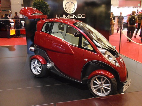 Red and white narrow tandem two-seater with a lifted tail on a dark stand under a LUMENEO sign. Opens a larger view