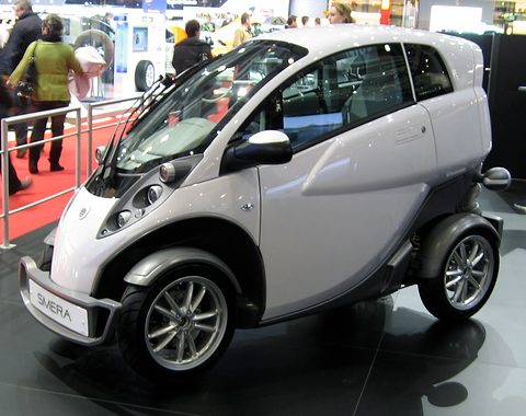 White and grey narrow two-seater with alloy wheels and a SMERA plate on a dark show floor, visitors behind. Opens a larger view