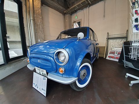 Blue rounded small saloon with whitewall tyres and a display placard, in a showroom with a dark wooden floor. Opens a larger view