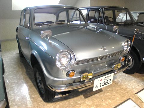 Silver-grey small saloon with chrome bumpers and amber lamps on a polished floor beside a black car.