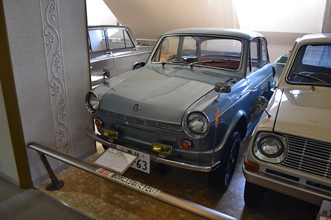 Pale blue-grey small saloon in a museum row behind a rail, a white car at right. Opens a larger view