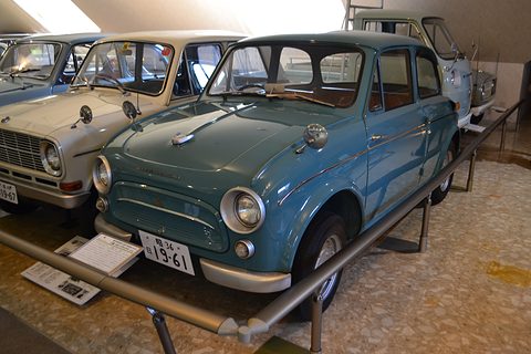 Blue small saloon behind a low steel rail in a museum row, a white car to its left. Opens a larger view