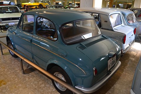 Rear three-quarter of a blue small saloon with an engine-lid grille, behind a steel rail. Opens a larger view