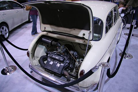 Rear lid raised on a cream small car, showing a compact black twin-cylinder engine with its cooling ducts. Opens a larger view