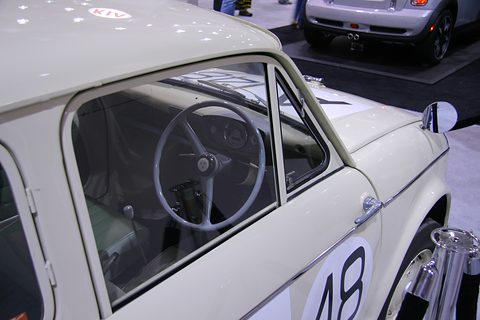 View through the side window of a cream car: a large thin two-spoke wheel and a plain painted dashboard. Opens a larger view