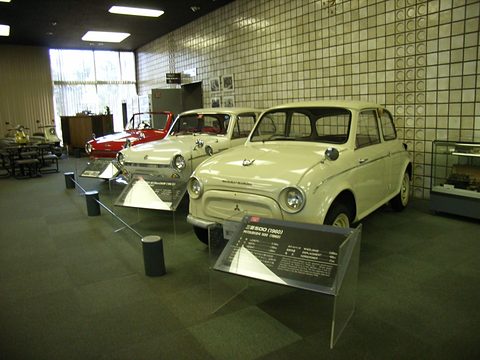Two cream small saloons on a dark floor in a tiled museum hall, with low display placards in front. Opens a larger view