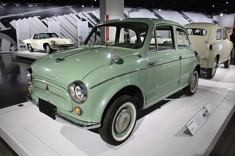 Pale green rounded small saloon on a white plinth, other small cars lined up behind in a bright gallery. Opens a larger view