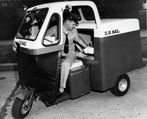 A uniformed postman seated in a small three-wheeled van lettered U.S. Mail, handing letters from its box.