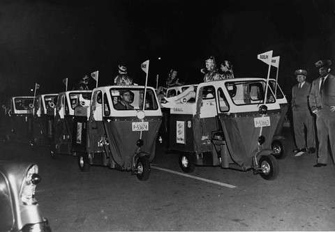 A row of small three-wheeled mail vans with pennants and drivers at night, officials standing alongside.