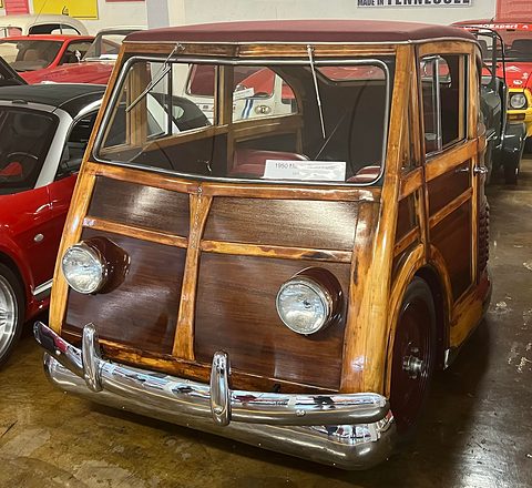 Front of a small varnished wooden estate car with twin round headlamps and a chrome bumper, other cars behind in a museum. Opens a larger view