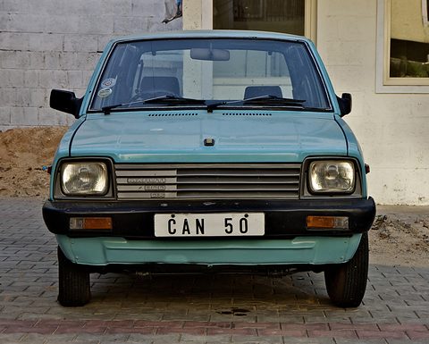 Front of a pale turquoise small hatchback with square headlamps, a slatted grille and a white plate, parked on brick paving.