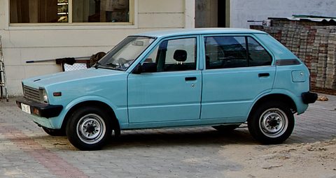 Side view of a pale turquoise small five-door hatchback with silver wheels, parked on paving in front of a white building. Opens a larger view