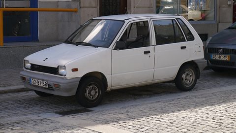 White small five-door hatchback with rectangular headlamps and a Portuguese plate, parked on a cobbled street by a shop. Opens a larger view