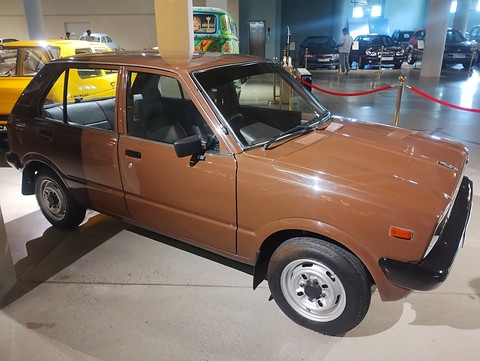 Brown small five-door hatchback with a black bumper on a shiny museum floor, a painted truck and yellow cars behind. Opens a larger view