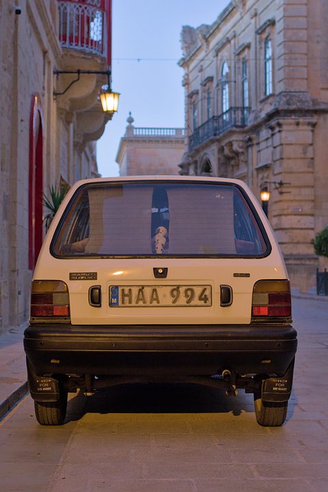 Rear view of a cream small hatchback with a white number plate, parked in a narrow lane between old stone buildings at dusk. Opens a larger view