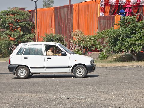 White small four-door hatchback in side view on a road, with an orange fence, bushes and street decorations behind. Opens a larger view
