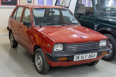 Red small five-door hatchback with square headlamps and an Indian plate, on a museum floor beside a dark green 4x4. Opens a larger view
