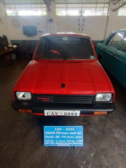 Head-on view of a red small hatchback above a blue information sign in a museum, other old cars along the walls. Opens a larger view