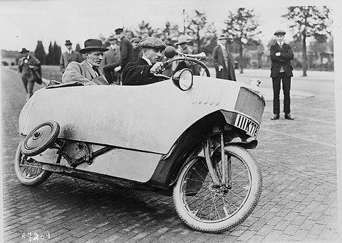 A pale, boat-shaped single-track car on a brick square, a raised outrigger wheel at its side and bystanders behind. Opens a larger view