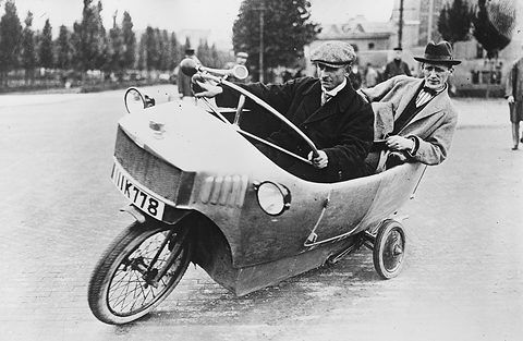 A narrow single-track car leaning into a turn on a paved square, a horn on its steering wheel and a small wheel at the rear.