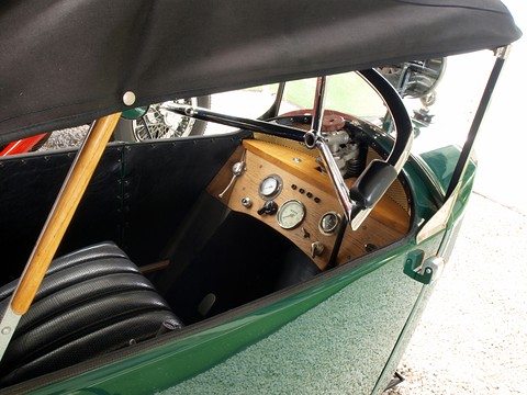Looking into a narrow cockpit: a wooden dashboard with round instruments, a steering wheel and pleated black seats. Opens a larger view
