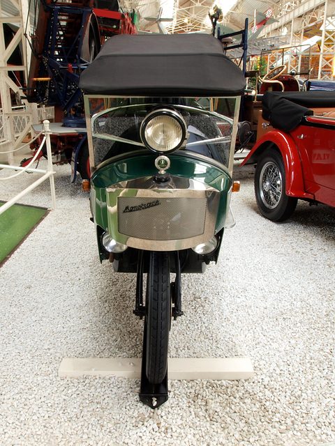 Front view of a very narrow green car with a single lamp above a mesh radiator and one front wheel beneath. Opens a larger view