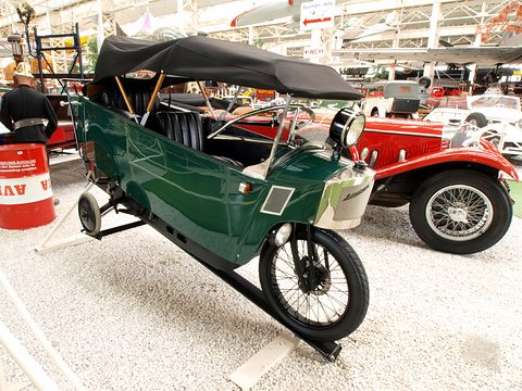 A long, narrow green single-track car with a black hood, a spoked front wheel and a small outrigger wheel at the rear. Opens a larger view