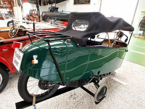 Rear three-quarter view of a green narrow car with a luggage rack on the tail, a black hood and an outrigger wheel lowered. Opens a larger view