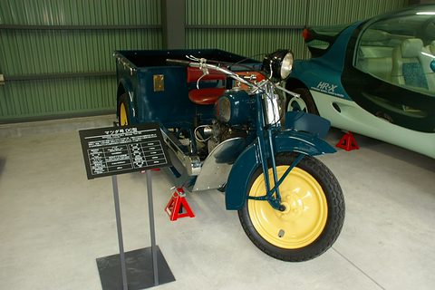 Teal three-wheeled truck with yellow wheels and a red seat, a museum sign on a stand in front of its engine. Opens a larger view