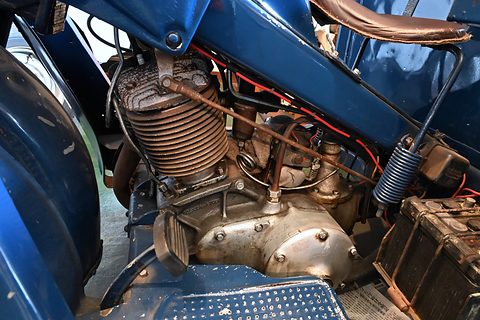 Finned single-cylinder engine under a blue frame, with a battery box and ribbed footboard beside it. Opens a larger view