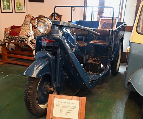 Blue three-wheeled truck with a step-through frame, a big headlamp and a museum label board in front. Opens a larger view