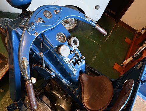 Blue curved handlebar frame with instruments, a gear lever and a sprung brown leather saddle.