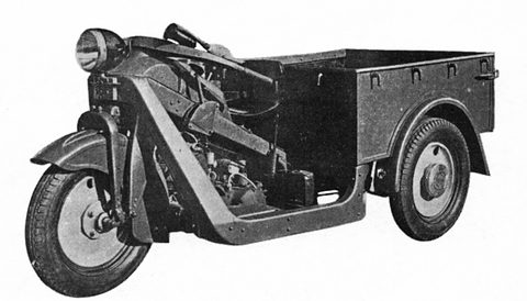 Black and white view of a three-wheeled truck with an open step-through frame, headlamp and cargo box.