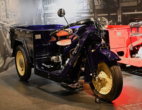 Dark blue three-wheeled truck with yellow wheels, mirrors and an open box, red display light behind. Opens a larger view