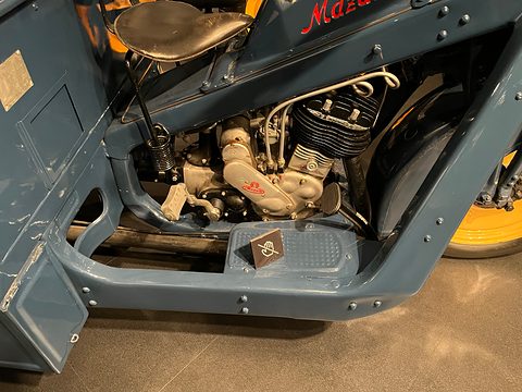 Single-cylinder engine with polished cases under a blue-grey frame and saddle, a ribbed footboard below. Opens a larger view
