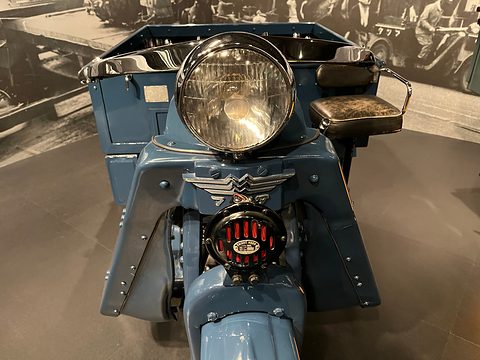 Big chrome headlamp, winged badge and a red horn grille on a blue-grey three-wheeler. Opens a larger view