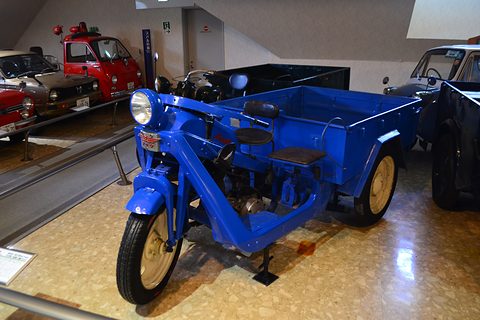 Bright blue three-wheeled truck with an open frame, small saddle and cargo bed, other old vehicles behind. Opens a larger view