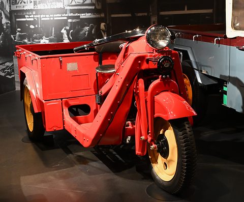 Bright red three-wheeled truck with cream wheels, a large headlamp and a step-through frame, a truck behind. Opens a larger view