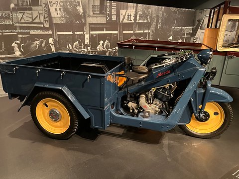 Side view of a blue-grey three-wheeled truck with yellow wheels, exposed engine and a flat cargo bed. Opens a larger view