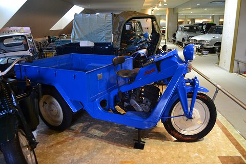Blue three-wheeler from the right showing its frame, engine and long cargo bed, cars displayed behind. Opens a larger view