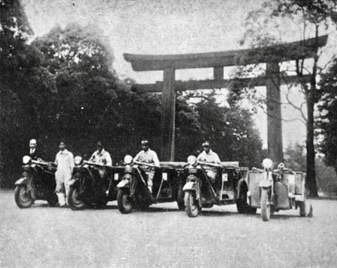 Five three-wheeled trucks parked in a row with their crews in white overalls beneath a large shrine gate.