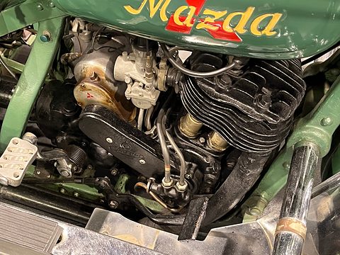 Finned single-cylinder engine with carburettor and pipes under a green tank lettered Mazda in gold script. Opens a larger view