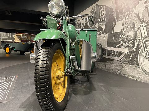 Front of a green three-wheeler: single front tyre in girder forks, one big headlamp and wide handlebars. Opens a larger view