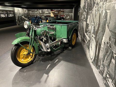 Green three-wheeler with yellow wheels, handlebars and an open cargo box, a black and white mural wall alongside. Opens a larger view