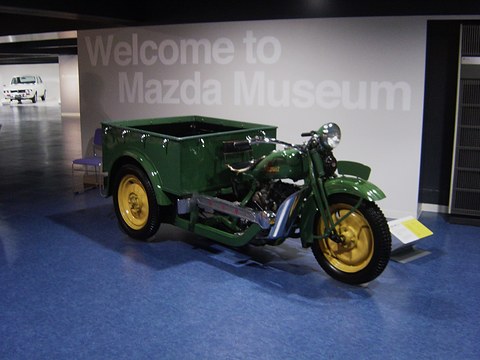 Green three-wheeled truck with yellow wheels on a blue floor below a large ‘Welcome to Mazda Museum’ sign. Opens a larger view
