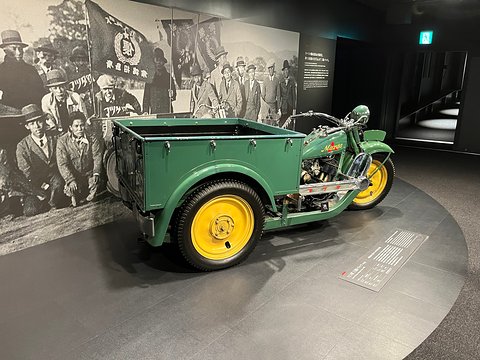 Rear three-quarter of a green three-wheeled truck with yellow wheels against a mural of a crowd. Opens a larger view