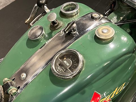 Top of a green fuel tank with a round speedometer, two filler caps, a switch and a black centre strip. Opens a larger view