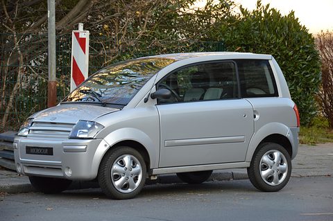 Small silver two-seat hatchback with a slatted nose parked at a kerb under bare branches. Opens a larger view