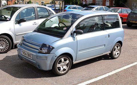 Pale blue small two-door microcar with a slatted nose parked among saloon cars. Opens a larger view